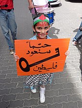 Das Bild zeigt ein junges Mädchen, das an einer Demonstration teilnimmt. Sie trägt ein Stirnband, das mit der palästinensischen Flagge verziert ist, und ihr Gesicht ist in den Farben Rot und Schwarz bemalt, die die Farben der Flagge widerspiegeln. Das Mädchen hält ein großes oranges Plakat mit arabischer Kalligrafie in Schwarz, die besagt: „Wir werden definitiv nach Palästina zurückkehren“. Das Schild zeigt das Bild eines Schlüssels (Viele Menschen, die 1948 vertrieben wurden oder geflohen sind, haben nur noch ihre Schlüssel von ihren alten Häusern, zu denen sie nicht zurückkehren können). Hinter ihr spielt ein anderer Junge, teilweise sichtbar, auf einer blauen Trommel. Der Hintergrund deutet darauf hin, dass dies in einem städtischen Umfeld stattfindet, mit einer grauen Kopfsteinpflasterstraße und Fahrzeugen in der Nähe.