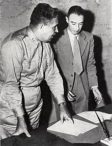 Photo en noir et blanc. Deux hommes debout, un habillé d'un uniforme, l'autre d'un complet, discutent près d'une table.