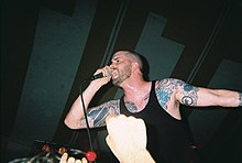Vocalist Andrew Schwab performs at a concert. He has shaved his head and is wearing a black sleeveless shirt that displays his tattoos.