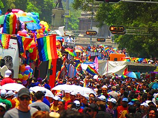 A crowd of people in a Pride March. Showing rainbow flags and rainbow colored ballons