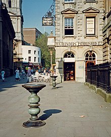 Two ornate metal pillars with large dishes on top in a paved street, with a eighteenth century stone building behind upon which can be seen the words "Tea Blenders Estabklishec 177-". People sitting at cafe style tables outside. On the right iron railings.