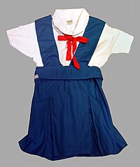 Una camicetta bianca a maniche corte con un ficco rosso a cui è abbinata una gonna-salopette blu Francia