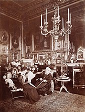 Photographie en noir et blanc d'une Victoria âgée assise à côté d'une jeune femme (Beatrice) lisant un journal. La pièce est richement décorée avec de nombreuses photographies et peintures et un large chandelier est suspendu au plafond.
