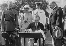 Coolidge assis à une table en extérieur signe un document en présence de plusieurs personnes.