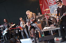 A rock band performing live on stage. A poster with the words "SPIN 20" is in the background.