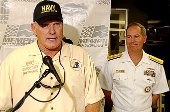 Un homme avec une casquette Navy parle dans un micro sous les yeux d'un militaire présent en arrière-plan.