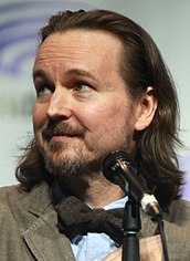 Zdjęcie reżysera Matta Reevesa w niebieskiej koszuli, brązowej marynarce i brązowej muszce, stojącego przed mikrofonem.