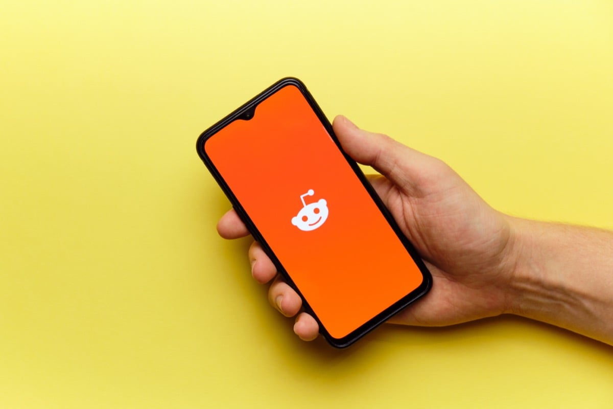 A hand holds a smartphone displaying the Reddit logo on an orange background
