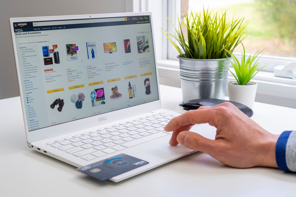 A person uses a laptop displaying the Amazon website to shop online, holding a credit card; the setting appears to be an outdoor cafe