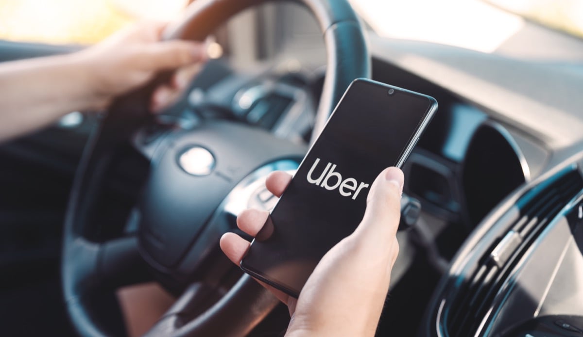 A smartphone displaying the Uber logo is held by a driver in a car; the driver is operating the vehicle